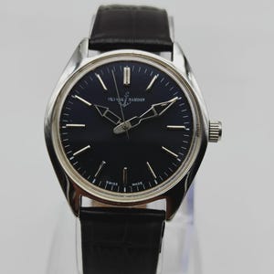 Puede incluir: Un reloj de pulsera Ulysse Nardin con esfera negra y agujas e indicadores plateados. El reloj tiene una caja plateada y una correa de cuero negro. El reloj se exhibe sobre un soporte transparente.