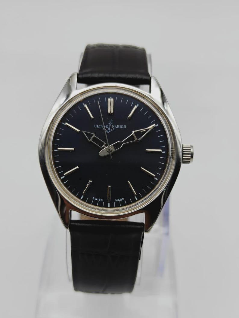 Puede incluir: Un reloj de pulsera Ulysse Nardin con esfera negra y agujas e indicadores plateados. El reloj tiene una caja plateada y una correa de cuero negro. El reloj se exhibe sobre un soporte transparente.