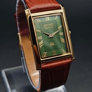 Puede incluir: Reloj Seiko Quartz rectangular dorado con esfera verde y n&uacute;meros romanos dorados. El reloj tiene agujas doradas y una correa de cuero marr&oacute;n. El reloj se muestra sobre un soporte transparente sobre un fondo oscuro.