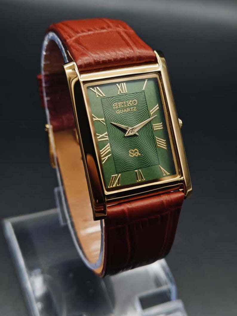 Puede incluir: Reloj Seiko Quartz rectangular dorado con esfera verde y n&uacute;meros romanos dorados. El reloj tiene agujas doradas y una correa de cuero marr&oacute;n. El reloj se muestra sobre un soporte transparente sobre un fondo oscuro.
