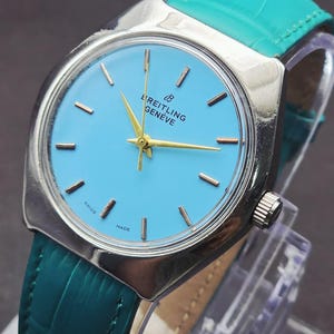 Könnte beinhalten: Eine Breitling Genève Armbanduhr mit hellblauem Zifferblatt, goldfarbenen Zeigern und goldfarbenen Stundenmarkierungen. Die Uhr hat ein silberfarbenes Metallgehäuse und ein strukturiertes blaugrünes Lederarmband. Breitling Genève und Swiss Made sind lesbar.