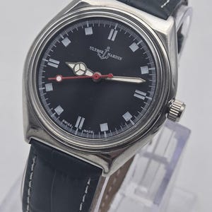 K&ouml;nnte beinhalten: Eine Ulysse Nardin-Uhr aus Edelstahl mit schwarzem Zifferblatt und dunkelblauem Lederarmband. Das Zifferblatt hat wei&szlig;e Markierungen und Zeiger mit einem roten Sekundenzeiger. Das Armband ist mit wei&szlig;em Faden gen&auml;ht.
