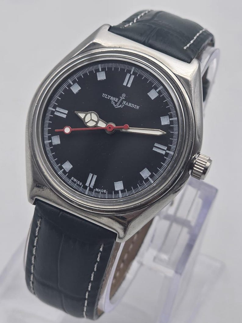 K&ouml;nnte beinhalten: Eine Ulysse Nardin-Uhr aus Edelstahl mit schwarzem Zifferblatt und dunkelblauem Lederarmband. Das Zifferblatt hat wei&szlig;e Markierungen und Zeiger mit einem roten Sekundenzeiger. Das Armband ist mit wei&szlig;em Faden gen&auml;ht.