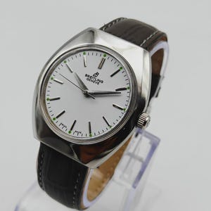 May include: A silver-toned Breitling Gen&egrave;ve watch with a white face, black hands, and green accents. The watch has a black leather band and is displayed on a clear stand. The watch face has the text "Breitling Gen&egrave;ve".