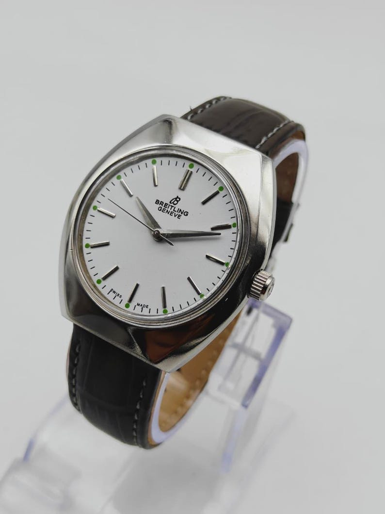 May include: A silver-toned Breitling Gen&egrave;ve watch with a white face, black hands, and green accents. The watch has a black leather band and is displayed on a clear stand. The watch face has the text "Breitling Gen&egrave;ve".