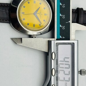 May include: A vintage wristwatch with a yellow face and silver case is measured with a digital caliper. The watch has a brown leather band. The caliper reads 40.23 mm, which is approximately 1.58 inches.