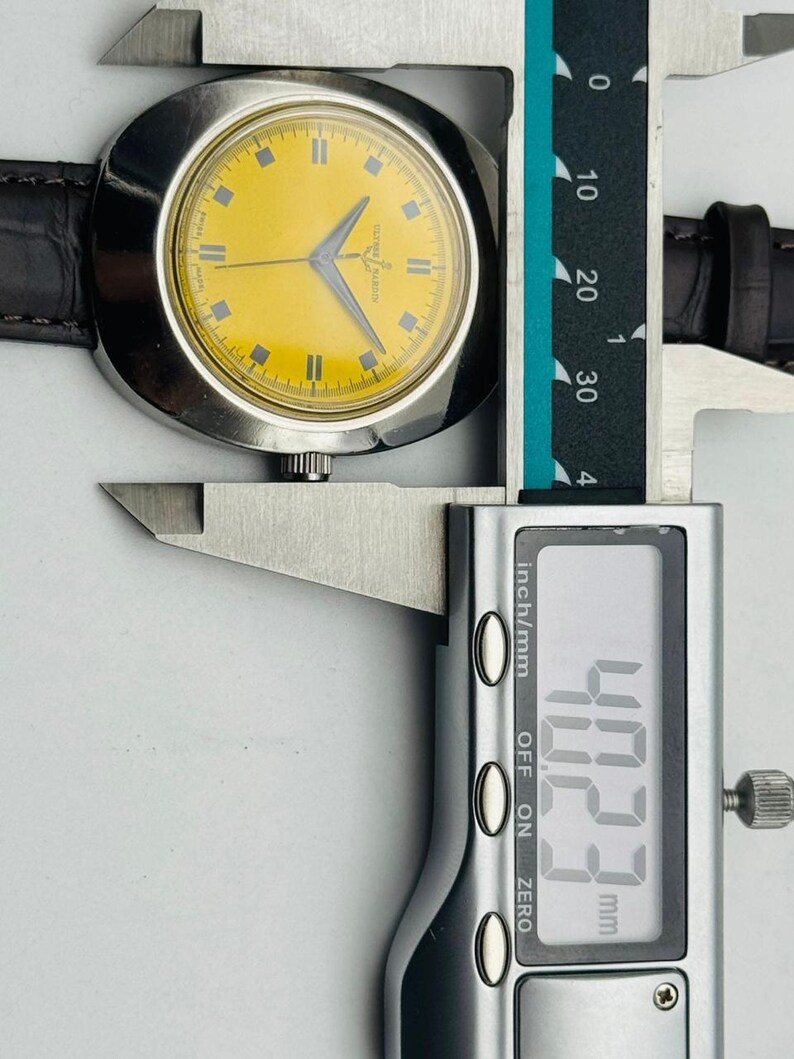 May include: A vintage wristwatch with a yellow face and silver case is measured with a digital caliper. The watch has a brown leather band. The caliper reads 40.23 mm, which is approximately 1.58 inches.