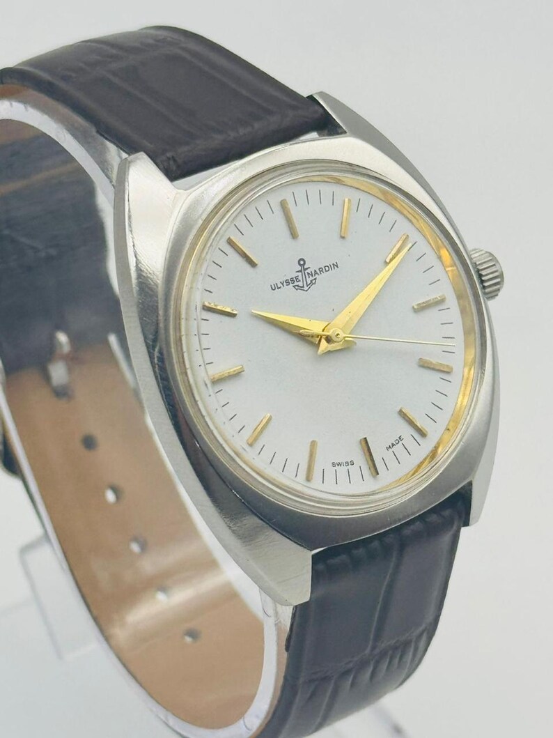 May include: A silver-toned wristwatch with a white face and gold hands. The watch features a black leather band and the brand name "Ulysse Nardin" on the face. The watch is a classic design.