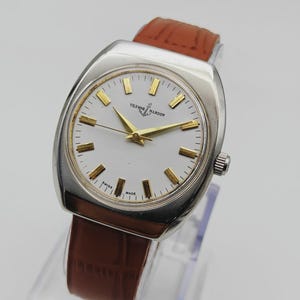 K&ouml;nnte beinhalten: Eine Vintage-Armbanduhr von Ulysse Nardin mit silberfarbenem Geh&auml;use und wei&szlig;em Zifferblatt. Die Uhr hat goldfarbene Zeiger und Stundenmarkierungen sowie ein braunes Lederarmband. Die Uhr ist mit "Swiss Made" gekennzeichnet.