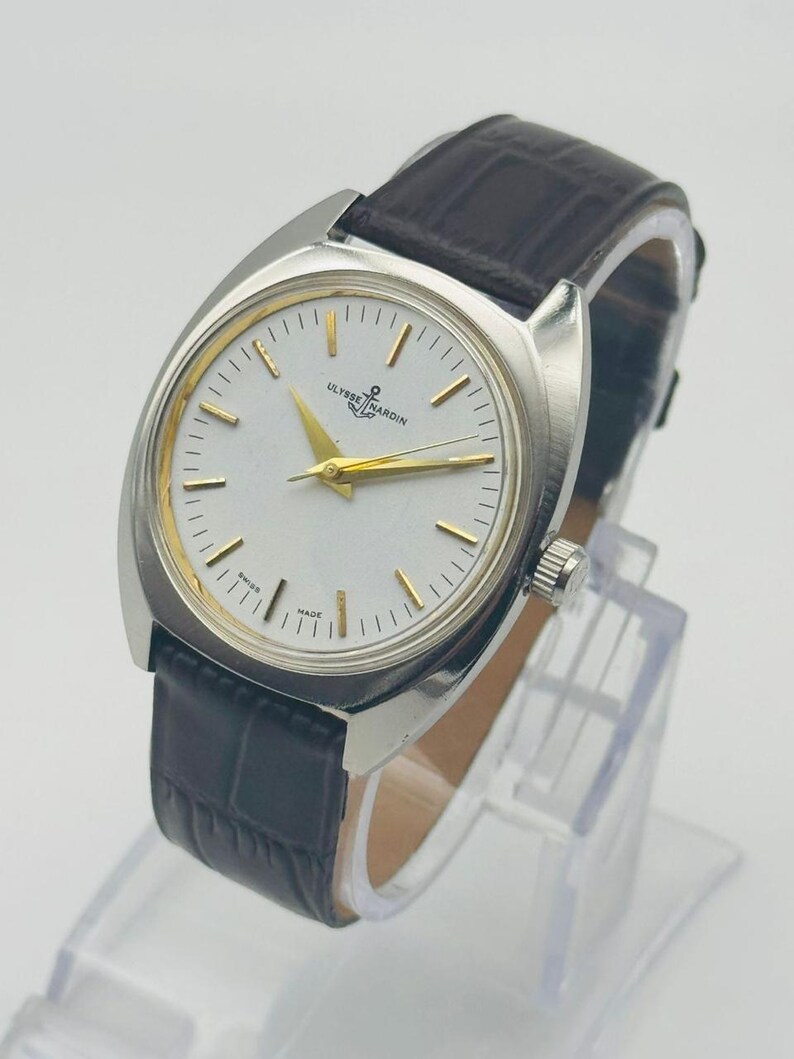 May include: A silver-toned Ulysse Nardin wristwatch with a white face and gold hands and markers. The watch has a dark brown leather band. The watch is displayed on a clear stand.