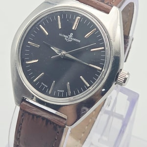 May include: A silver-toned Ulysse Nardin wristwatch with a black face and brown leather band. The watch face has gold-colored hour markers and hands. The band is a dark brown color with a textured appearance. The watch is labeled "SWISS MADE".