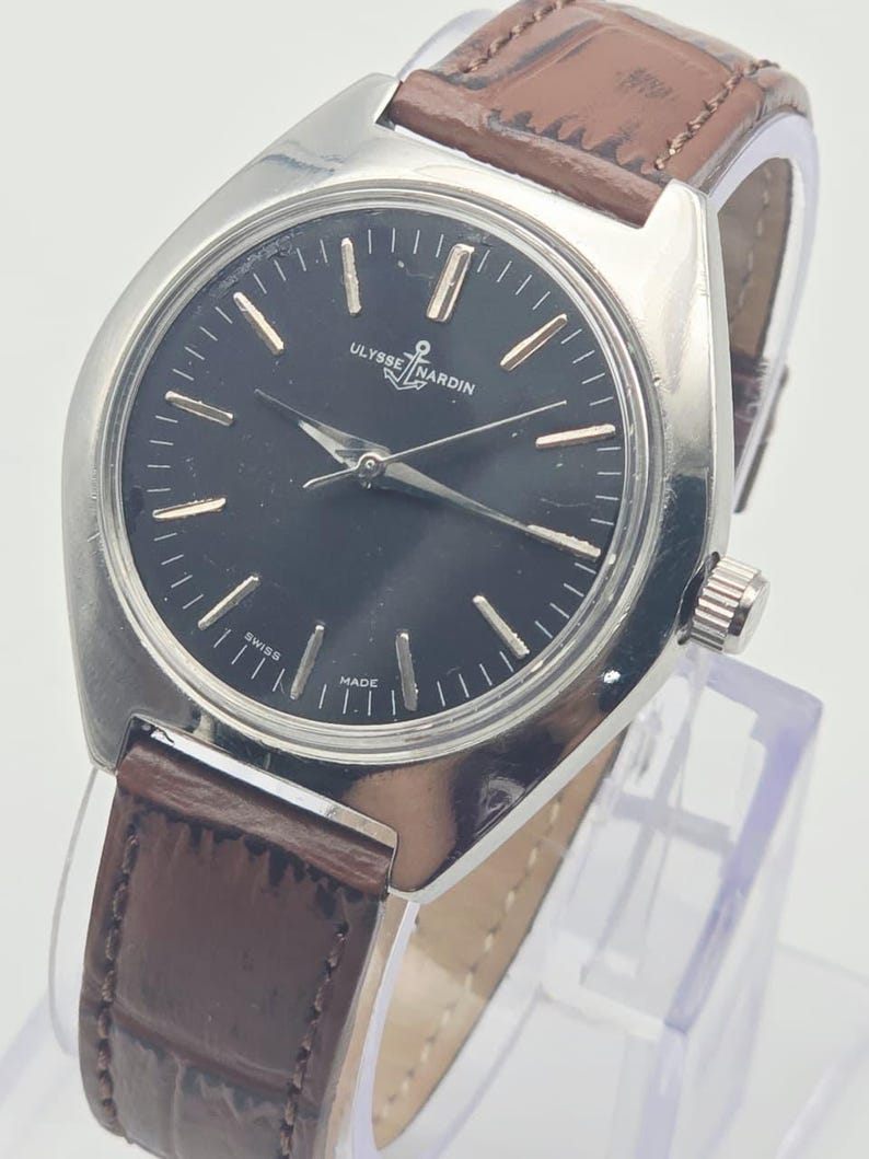 May include: A silver-toned Ulysse Nardin wristwatch with a black face and brown leather band. The watch face has gold-colored hour markers and hands. The band is a dark brown color with a textured appearance. The watch is labeled "SWISS MADE".