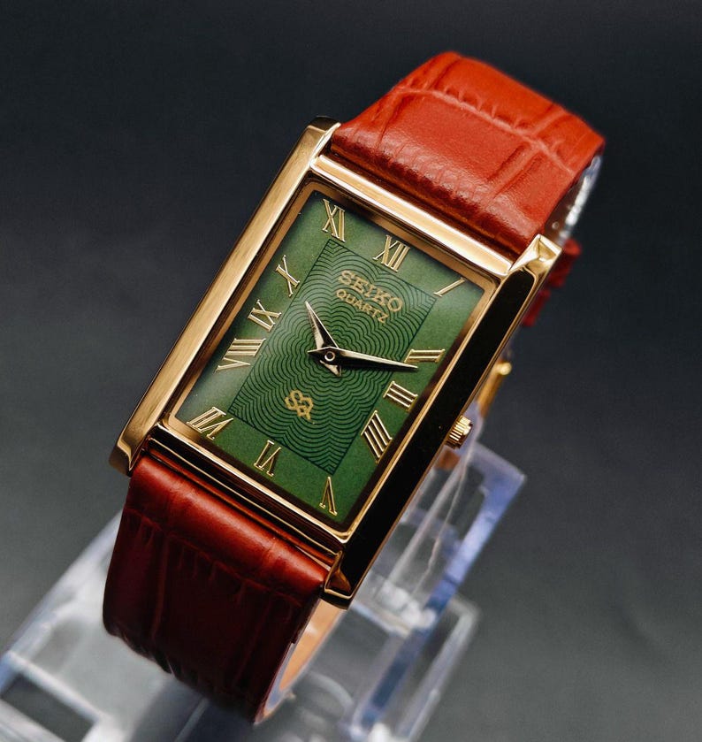 Puede incluir: Un reloj Seiko Quartz dorado con una esfera verde rectangular y n&uacute;meros romanos. El reloj tiene una correa de cuero marr&oacute;n. La esfera del reloj tiene un patr&oacute;n texturizado y el logotipo de Seiko Quartz.