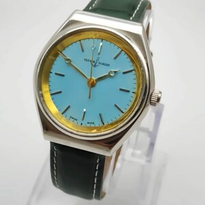 May include: A silver Ulysse Nardin watch with a light blue face and gold hands and accents. The watch has a green leather strap with white stitching. The watch face has the text "Ulysse Nardin" and "Swiss Made".