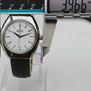May include: A silver Breitling watch with a white face and black leather band is measured with a digital caliper. The watch face has black hands and green accents. The caliper reads 39.66 mm, which is approximately 1.56 inches.