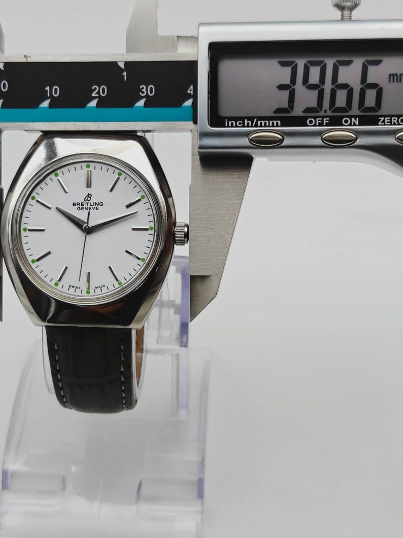 May include: A silver Breitling watch with a white face and black leather band is measured with a digital caliper. The watch face has black hands and green accents. The caliper reads 39.66 mm, which is approximately 1.56 inches.