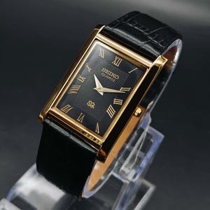 May include: A rectangular Seiko Quartz watch with a black face and gold-colored frame. The watch features Roman numeral hour markers and a black leather band. The watch is displayed on a clear stand against a dark background.