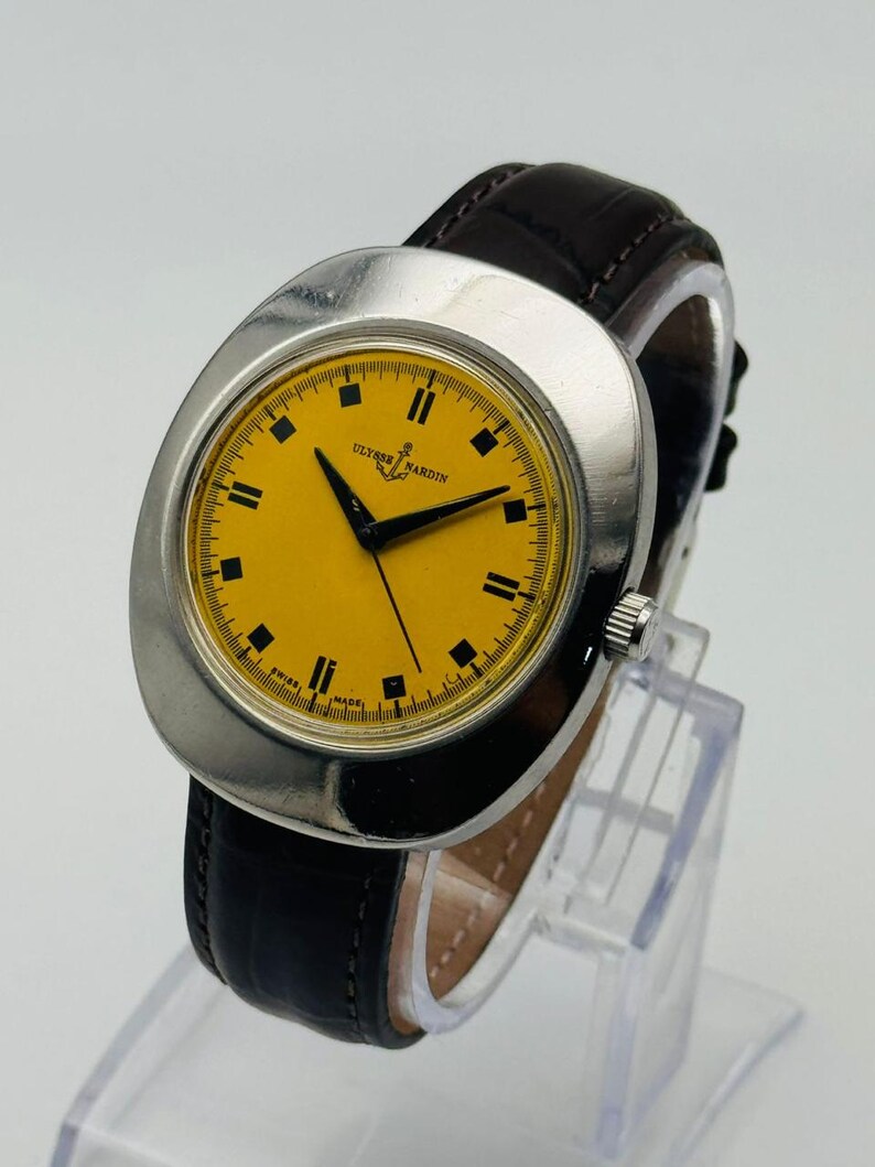 May include: A vintage Ulysse Nardin wristwatch with a bright yellow face and black hands. The watch has a silver-colored case and a dark brown leather band. The watch face has black square hour markers and the words "Ulysse Nardin" are printed on the face.