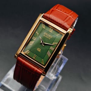 Puede incluir: Un reloj Seiko Quartz dorado con una esfera verde rectangular y n&uacute;meros romanos. El reloj tiene una correa de cuero marr&oacute;n. La esfera del reloj tiene un patr&oacute;n texturizado y el logotipo de Seiko Quartz.