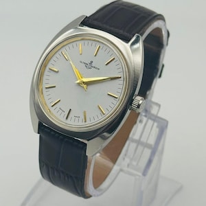 May include: A silver-toned Ulysse Nardin wristwatch with a white face, gold hands, and a black leather band. The watch has a rectangular case and a textured band. The watch is displayed on a clear stand.