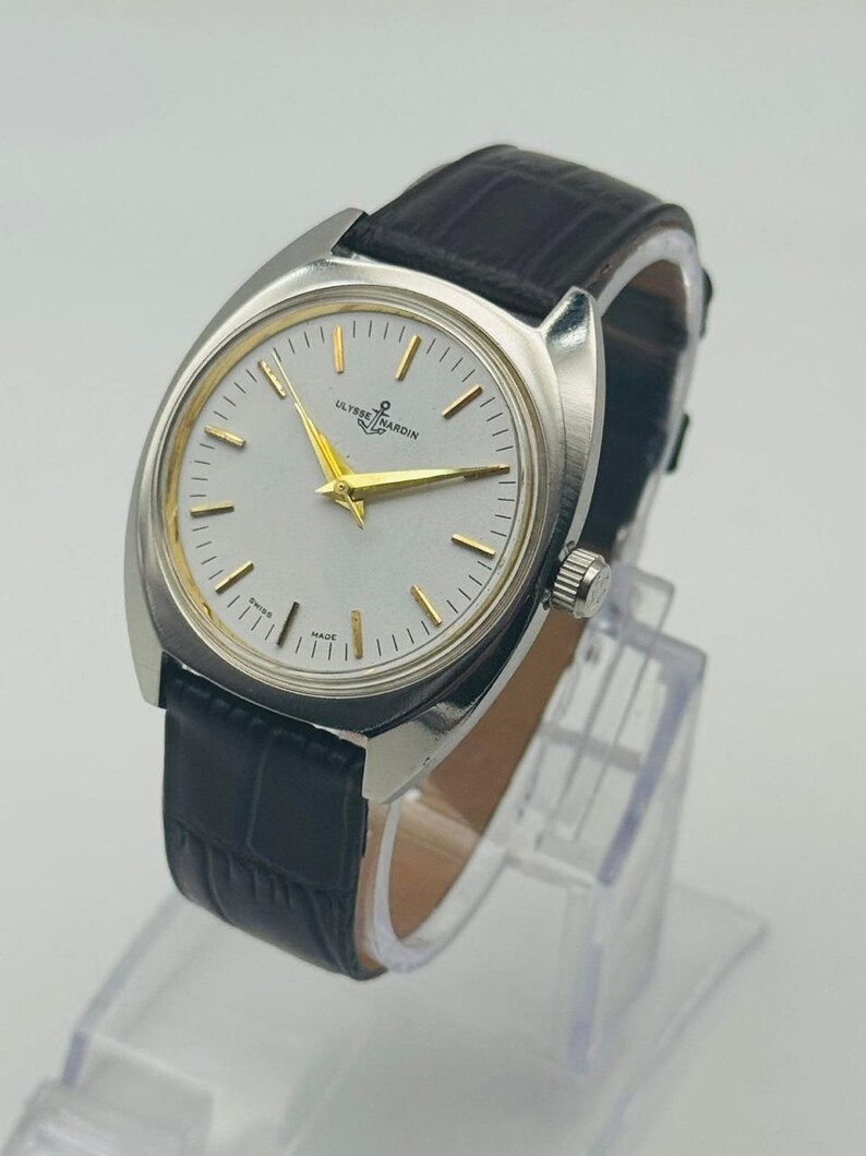 May include: A silver-toned Ulysse Nardin wristwatch with a white face, gold hands, and a black leather band. The watch has a rectangular case and a textured band. The watch is displayed on a clear stand.