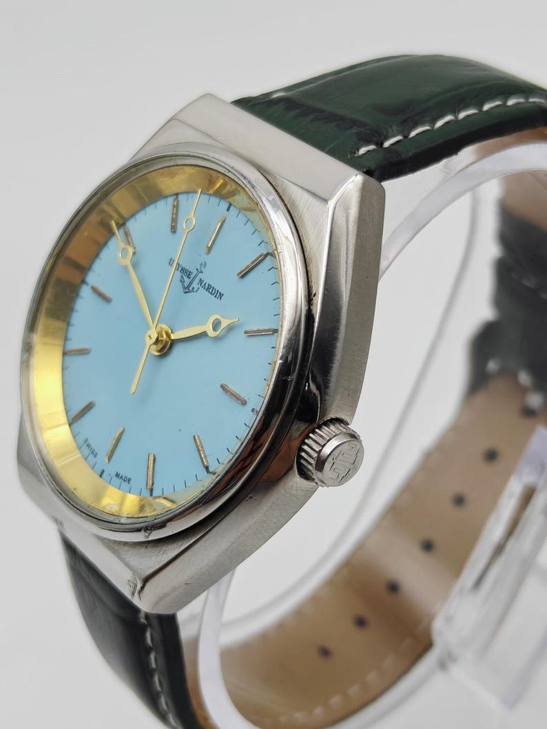 May include: A silver Ulysse Nardin wristwatch with a light blue face and gold hands. The watch features a black leather strap with white stitching. The watch face displays the brand name and an anchor logo.