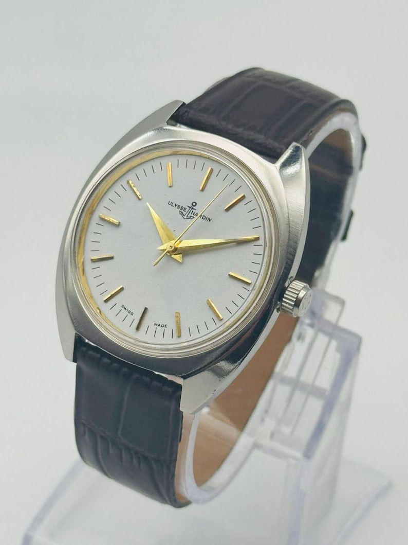 May include: A silver-toned wristwatch with a light-colored face and gold-colored hands and markers. The watch features the text "ULYSSE NARDIN" and a dark brown leather band. The watch is displayed on a clear stand.