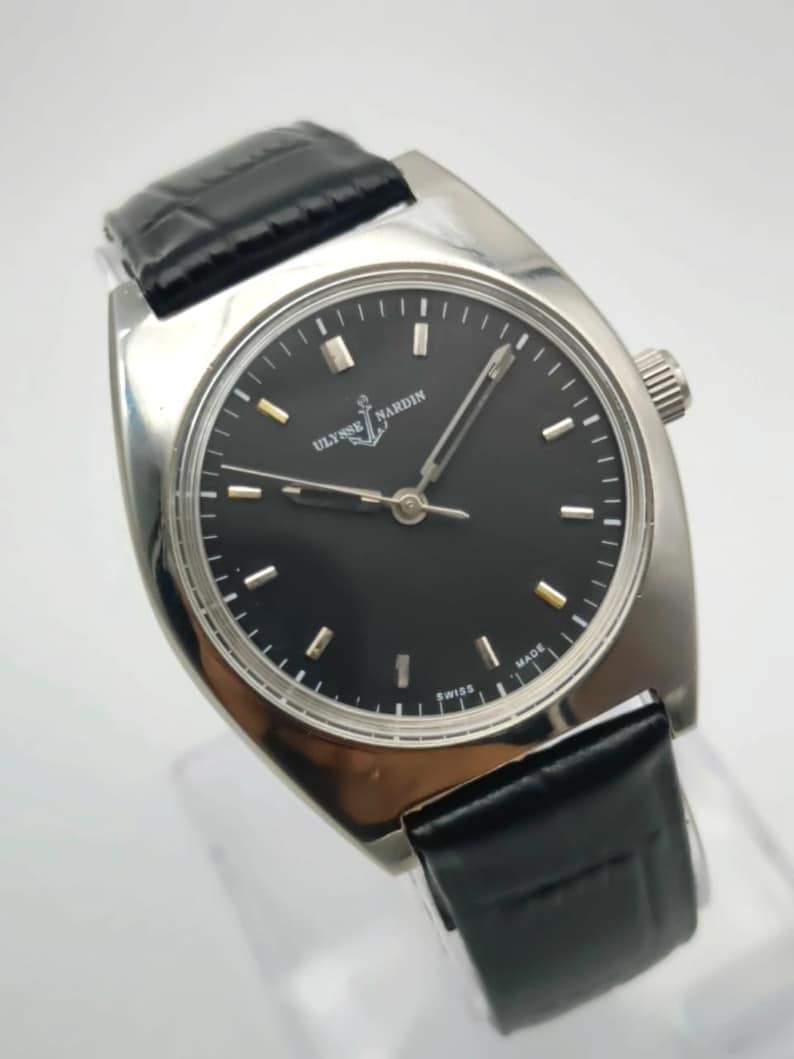 May include: A silver-toned wristwatch with a black face and a black leather band. The watch face features the brand name "ULYSSE NARDIN" and the word "SWISS MADE". The watch has a rectangular case and a crown on the side.