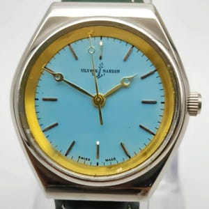 May include: A silver Ulysse Nardin wristwatch with a light blue face and gold accents. The watch features gold hands and a green leather strap. The words "SWISS MADE" are visible on the watch face.