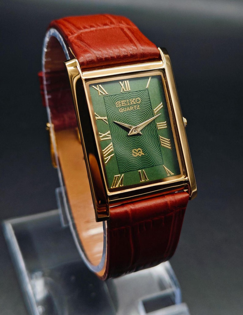 Puede incluir: Un reloj Seiko Quartz dorado con una esfera verde rectangular y n&uacute;meros romanos dorados. El reloj tiene una correa de cuero marr&oacute;n. La esfera del reloj tiene el logotipo de Seiko Quartz y un patr&oacute;n texturizado. El reloj se exhibe sobre un soporte transparente.