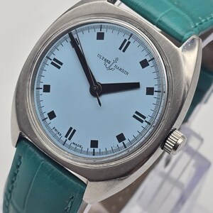 Pu&ograve; includere: Un orologio da polso argentato con quadrante azzurro e lancette e indici neri. L'orologio ha un cinturino in pelle verde. Sul quadrante dell'orologio sono impresse le parole "Ulysse Nardin" e "Swiss Made".