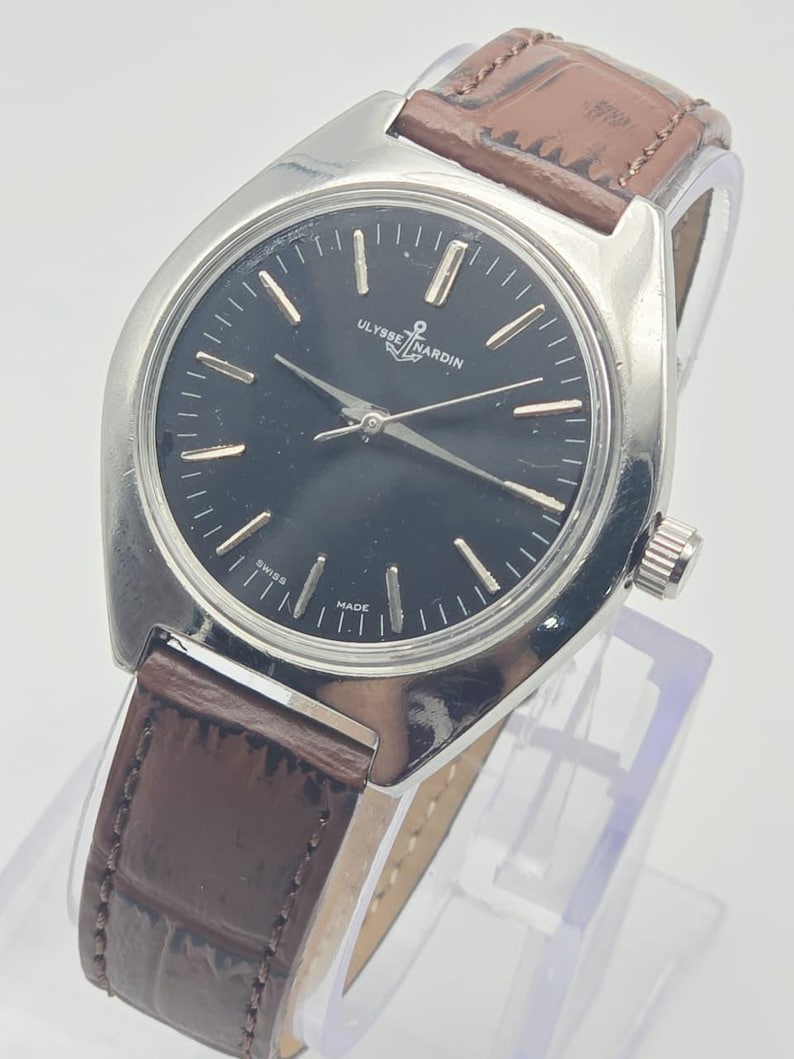 May include: A silver-toned Ulysse Nardin wristwatch with a black face and brown leather band. The watch has silver hands and markers, and the band is a dark brown with a textured pattern. The watch face reads "Ulysse Nardin" and "Swiss Made."
