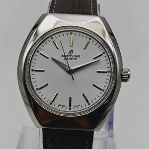 May include: A Breitling Geneve watch with a silver case and a white face. The watch has a dark brown leather band and a silver crown. The face features black hands, black markers, and green dots. The text "SWISS MADE" is visible.