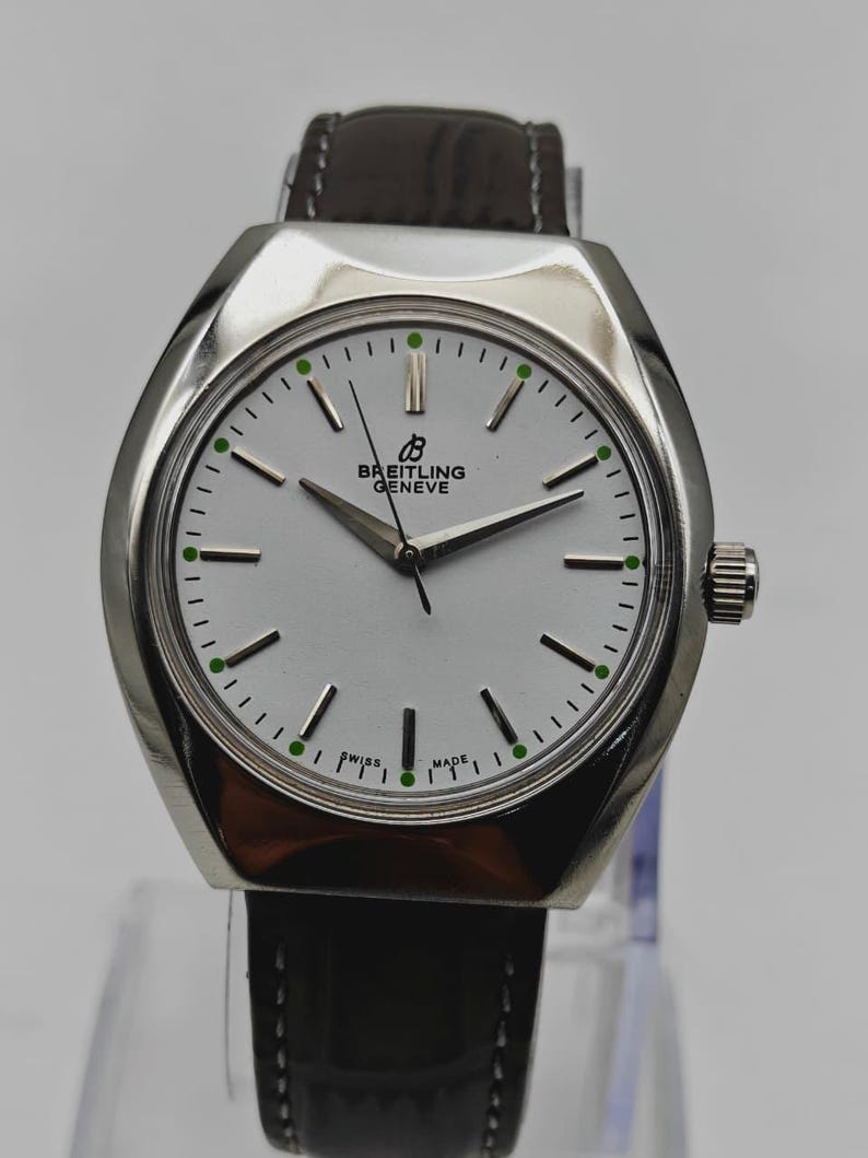 May include: A Breitling Geneve watch with a silver case and a white face. The watch has a dark brown leather band and a silver crown. The face features black hands, black markers, and green dots. The text "SWISS MADE" is visible.
