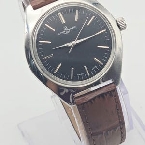 May include: A silver-toned wristwatch with a black face and brown leather band. The watch face features silver hour markers and hands, with the brand name "ULYSSE NARDIN" and an anchor logo. The band is approximately 0.75 inches wide.