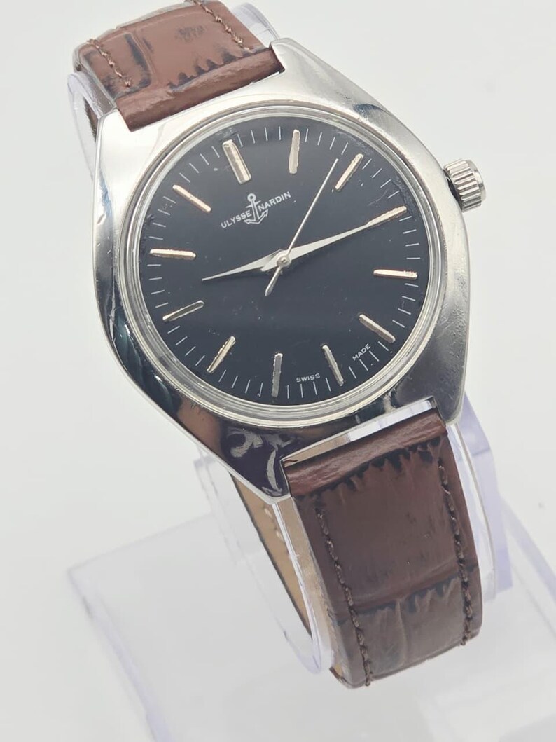 May include: A silver-toned wristwatch with a black face and brown leather band. The watch face features silver hour markers and hands, with the brand name "ULYSSE NARDIN" and an anchor logo. The band is approximately 0.75 inches wide.