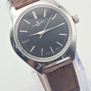 May include: A silver-toned Ulysse Nardin wristwatch with a black face and brown leather band. The watch face features silver hour markers and hands, with the Ulysse Nardin logo and "SWISS MADE" text. The band has a textured, crocodile-like pattern.