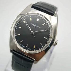 May include: A silver-toned Ulysse Nardin wristwatch with a black dial and a black leather band. The watch has a rectangular face with silver hour markers and hands. The watch is marked "Swiss Made".