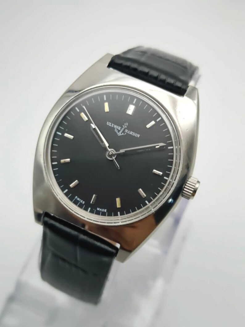 May include: A silver-toned Ulysse Nardin wristwatch with a black dial and a black leather band. The watch has a rectangular face with silver hour markers and hands. The watch is marked "Swiss Made".