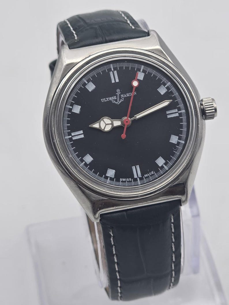 K&ouml;nnte beinhalten: Eine Ulysse Nardin-Uhr aus Edelstahl mit schwarzem Zifferblatt, wei&szlig;en Markierungen und einem roten Sekundenzeiger. Die Uhr hat ein Lederarmband mit wei&szlig;en N&auml;hten. Die Uhr wird auf einem transparenten St&auml;nder pr&auml;sentiert.