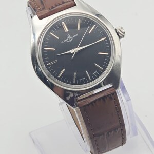 May include: A silver-toned wristwatch with a black face and brown leather band. The watch features silver hands and markers, with the brand name "ULYSSE NARDIN" displayed. The watch is displayed on a clear stand.