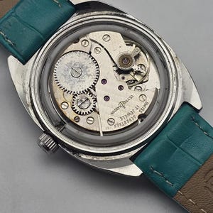Pu&ograve; includere: Un orologio da polso vintage con il retro aperto, che rivela i meccanismi interni. La cassa argentata circonda gli ingranaggi, le molle e i gioielli del meccanismo. Il cinturino &egrave; in pelle turchese.