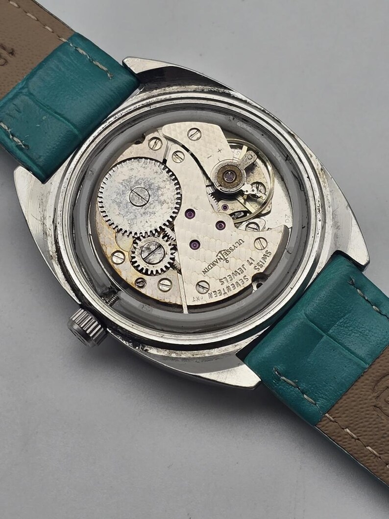 Pu&ograve; includere: Un orologio da polso vintage con il retro aperto, che rivela i meccanismi interni. La cassa argentata circonda gli ingranaggi, le molle e i gioielli del meccanismo. Il cinturino &egrave; in pelle turchese.