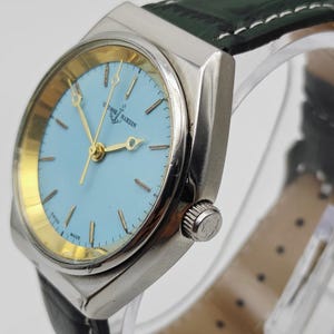 May include: A silver Ulysse Nardin wristwatch featuring a light blue face with gold hands and markers. The watch has a black leather strap with white stitching. The watch face displays the brand name and an anchor logo. The watch is a classic design.