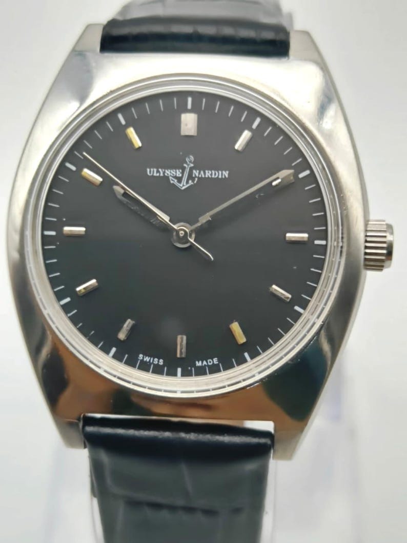 May include: A silver-toned Ulysse Nardin wristwatch with a black face and a black leather band. The watch face has silver and gold-toned markers and hands. The watch is marked "Swiss Made".
