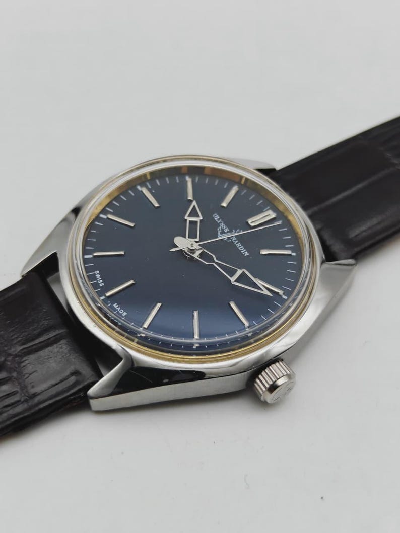 Puede incluir: Un reloj de pulsera plateado con esfera azul oscuro y detalles dorados. El reloj presenta agujas e indicadores plateados, una correa de cuero negro texturizado y una corona en el lateral. La esfera del reloj tiene el texto "Ulysse Nardin" y "Swiss Made".