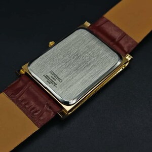 Puede incluir: Un reloj Seiko rectangular dorado con una correa de cuero marr&oacute;n. La parte trasera del reloj es plateada con el logotipo y el texto de Seiko. La correa es de color marr&oacute;n claro en la parte inferior y marr&oacute;n oscuro texturizado en la parte superior.