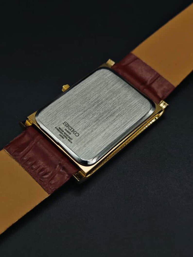 Puede incluir: Un reloj Seiko rectangular dorado con una correa de cuero marr&oacute;n. La parte trasera del reloj es plateada con el logotipo y el texto de Seiko. La correa es de color marr&oacute;n claro en la parte inferior y marr&oacute;n oscuro texturizado en la parte superior.