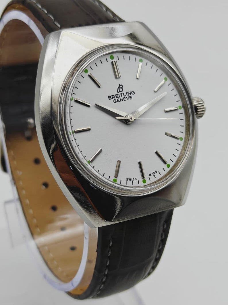 May include: A silver Breitling Geneve watch with a white face, black leather band, and green accents. The watch features a round face with the brand name and the words "SWISS MADE". The watch band is approximately 8 inches long.