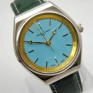 May include: A silver Ulysse Nardin watch with a light blue face and gold-coloured accents. The watch has a dark green leather strap. The watch face has black hour markers and hands. The watch is Swiss made.